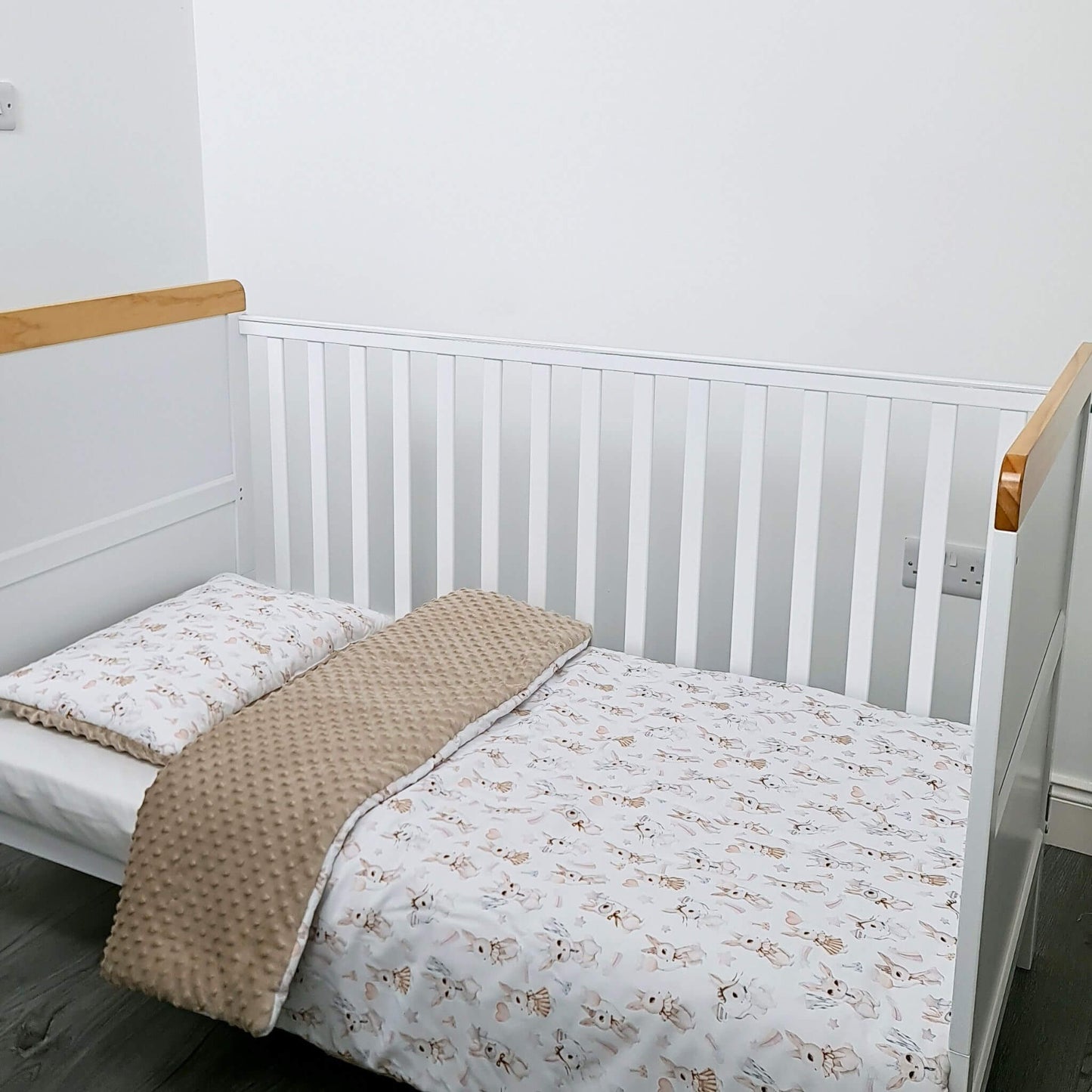 Crib with white bedding and a beige blanket in a room with a white wall and wooden floor. toddler size quilt with bunnies pattern