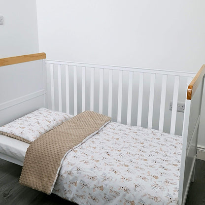 Crib with white bedding and a beige blanket in a room with a white wall and wooden floor. toddler size quilt with bunnies pattern