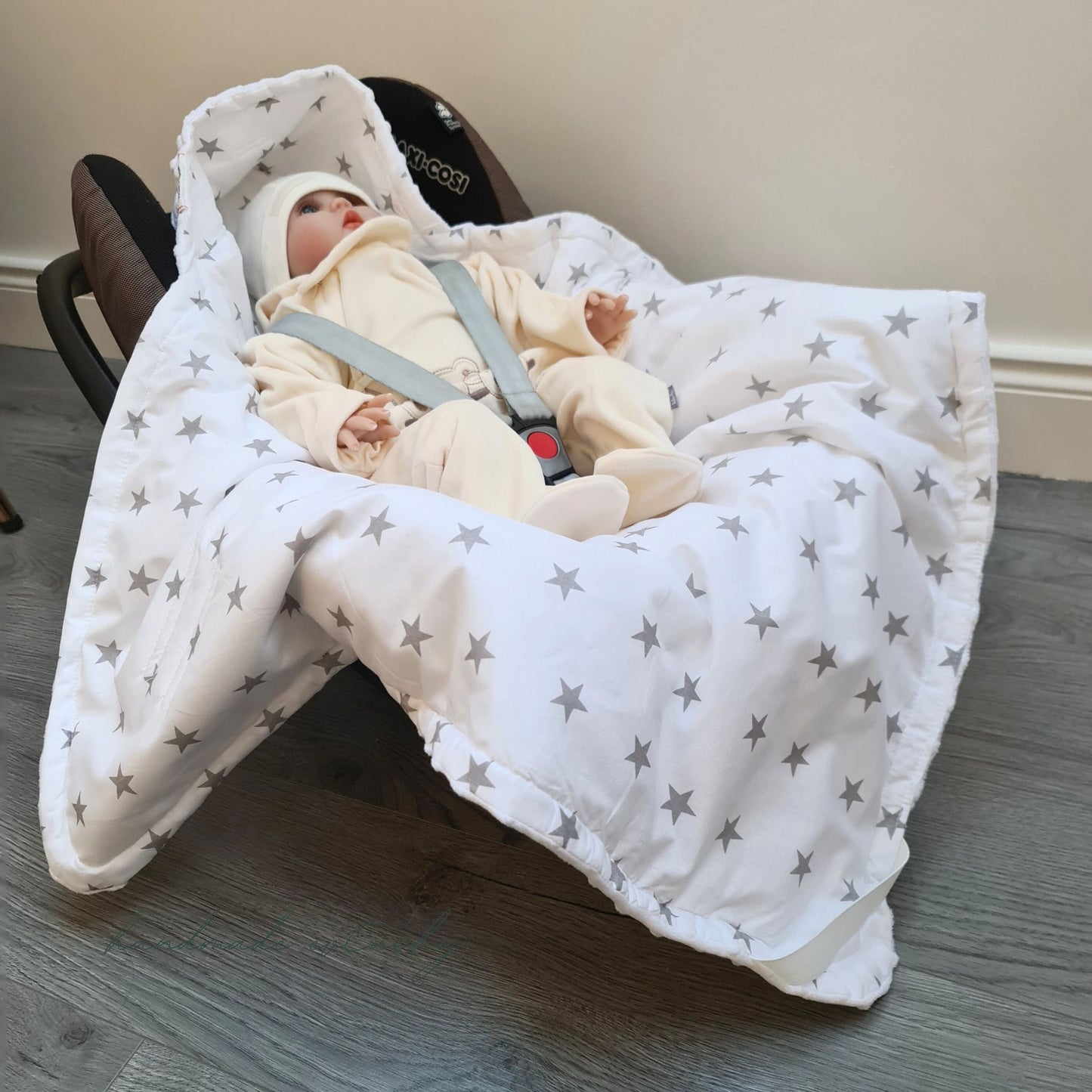 Image: A white car seat blanket with grey stars, featuring a convenient hood for added comfort and warmth.